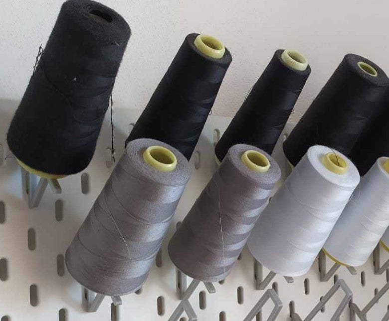 An einer weißen Skadis Lochwand sind mehrere Halter montiert, die große Garnrollen für Overlock-Nähmaschinen in verschiedenen Farben (Schwarz, Grau, Weiß) ordentlich und griffbereit halten.