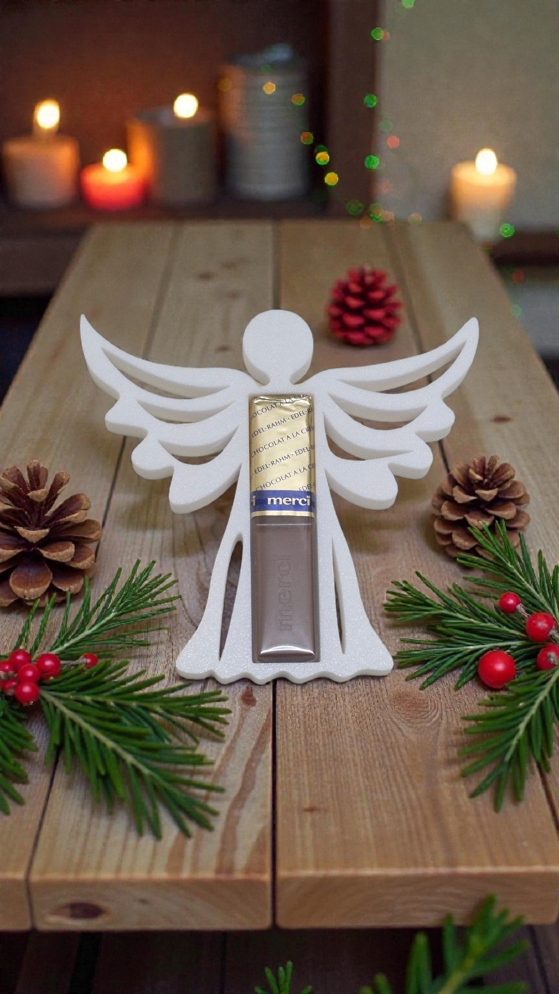 Weißer Engel Schokoladen-Ständer mit Merci Riegel, umgeben von Tannenzweigen, Beeren und Tannenzapfen als festliche Weihnachtsdeko.