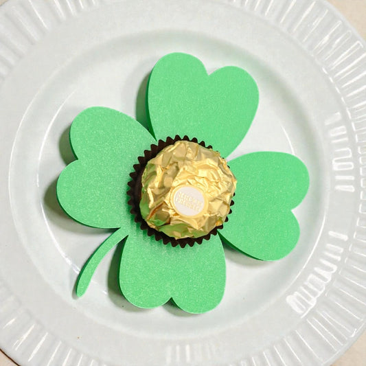Glücksklee Pralinen-Halter mit goldener Praline, präsentiert auf einem weißen, strukturierten Teller als Gastgeschenk-Idee.